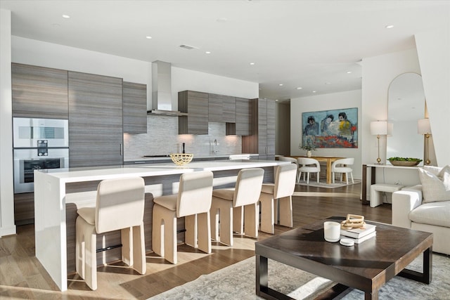 kitchen featuring light wood-type flooring, a kitchen breakfast bar, a kitchen island, wall chimney exhaust hood, and tasteful backsplash