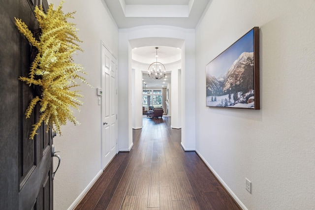 hallway featuring a tray ceiling, dark wood-style flooring, arched walkways, an inviting chandelier, and baseboards