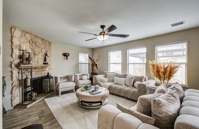 living room featuring ceiling fan, wood-type flooring, a stone fireplace, and a textured ceiling