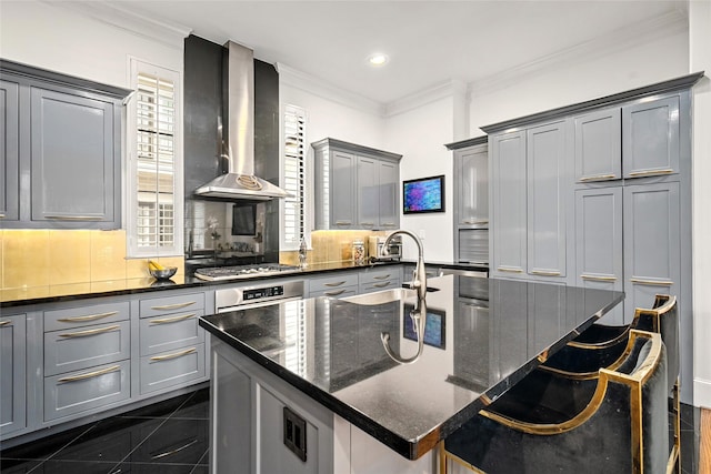 kitchen featuring a center island with sink, stainless steel gas cooktop, dark stone countertops, wall chimney range hood, and a breakfast bar area