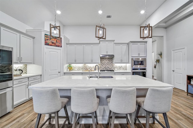 kitchen featuring a center island with sink, sink, light stone counters, stainless steel microwave, and pendant lighting