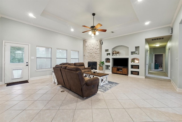 tiled living room with built in shelves, a stone fireplace, ceiling fan, a raised ceiling, and crown molding