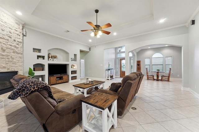 living room featuring light tile patterned floors, a stone fireplace, a raised ceiling, built in features, and ornamental molding