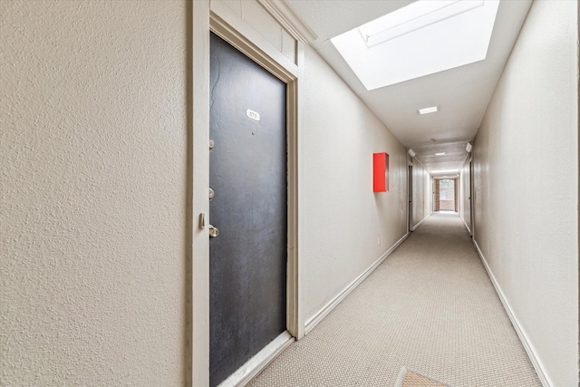 hallway featuring a skylight, a textured wall, baseboards, and light colored carpet