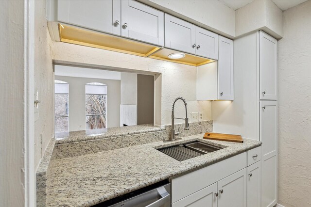 kitchen featuring white cabinets, dishwasher, a textured wall, light stone countertops, and a sink