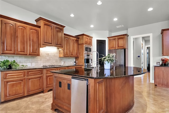 kitchen featuring appliances with stainless steel finishes, backsplash, sink, a kitchen island with sink, and dark stone countertops