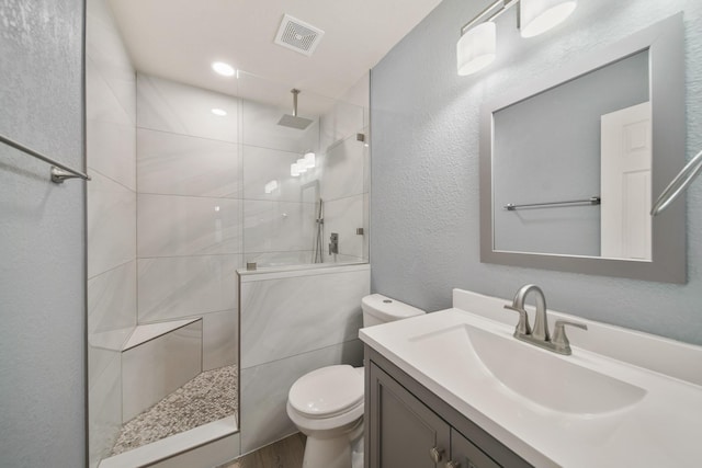 bathroom featuring a walk in shower, a textured wall, toilet, vanity, and visible vents