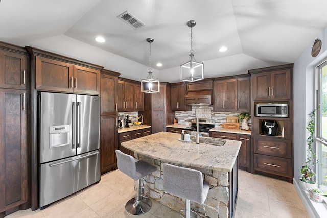 kitchen with appliances with stainless steel finishes, a center island with sink, hanging light fixtures, light stone counters, and vaulted ceiling