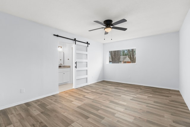 unfurnished bedroom featuring sink, light wood-type flooring, ensuite bathroom, ceiling fan, and a barn door