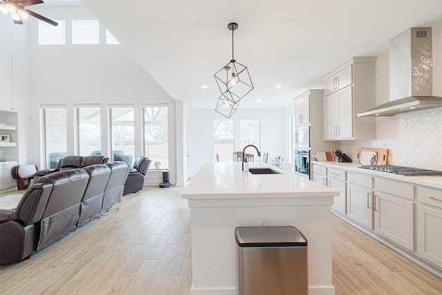 kitchen featuring a center island with sink, sink, wall chimney range hood, white cabinets, and hanging light fixtures