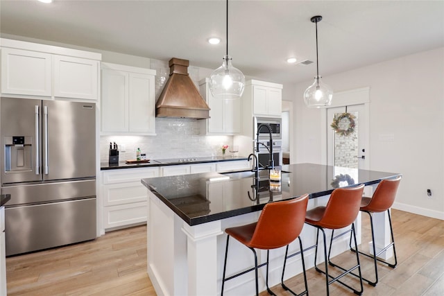 kitchen with a kitchen island with sink, custom exhaust hood, stainless steel appliances, white cabinetry, and a sink