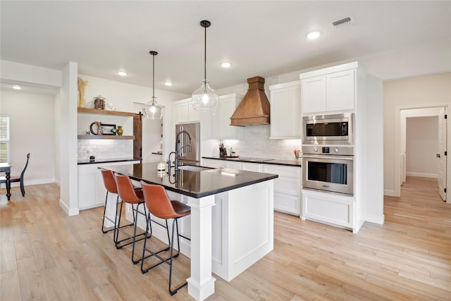 kitchen with dark countertops, custom range hood, a kitchen island with sink, stainless steel appliances, and open shelves