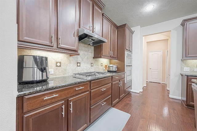 kitchen featuring dark wood-type flooring, stainless steel appliances, a textured ceiling, dark stone counters, and backsplash