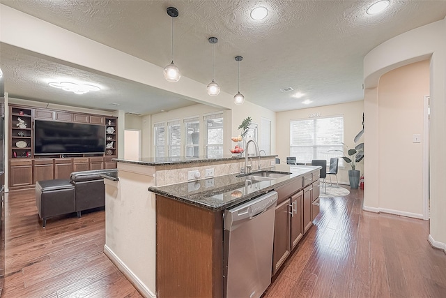 kitchen featuring hanging light fixtures, an island with sink, stainless steel dishwasher, sink, and dark stone countertops