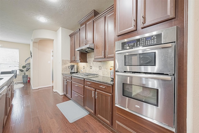 kitchen featuring appliances with stainless steel finishes, dark wood-type flooring, a textured ceiling, dark stone counters, and backsplash