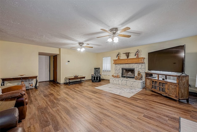 living room featuring a ceiling fan, a stone fireplace, a textured ceiling, wood finished floors, and baseboards