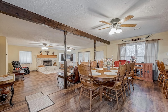 dining area with visible vents, wood finished floors, a textured ceiling, a stone fireplace, and beam ceiling