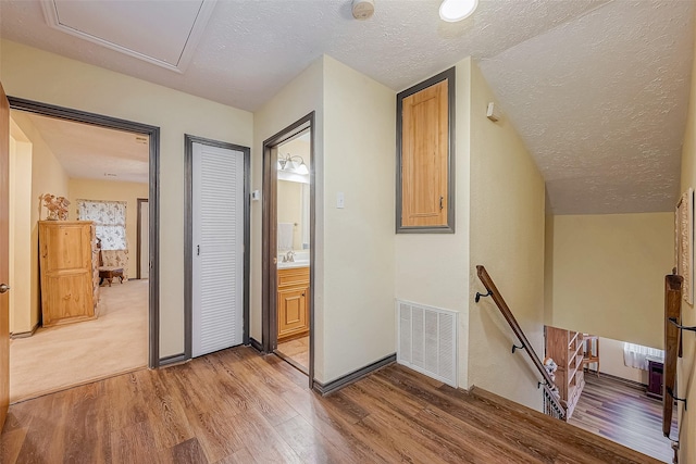 corridor featuring light wood-style floors, visible vents, a textured ceiling, and an upstairs landing