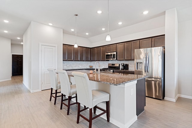 kitchen with an island with sink, stainless steel appliances, decorative light fixtures, tasteful backsplash, and dark stone countertops