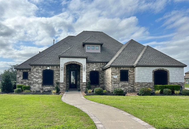 french country home featuring a shingled roof, a front yard, and brick siding