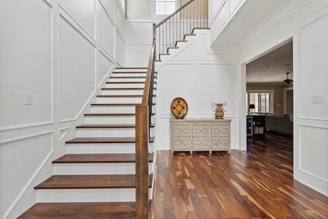 stairway featuring a ceiling fan, a towering ceiling, wood finished floors, crown molding, and a decorative wall