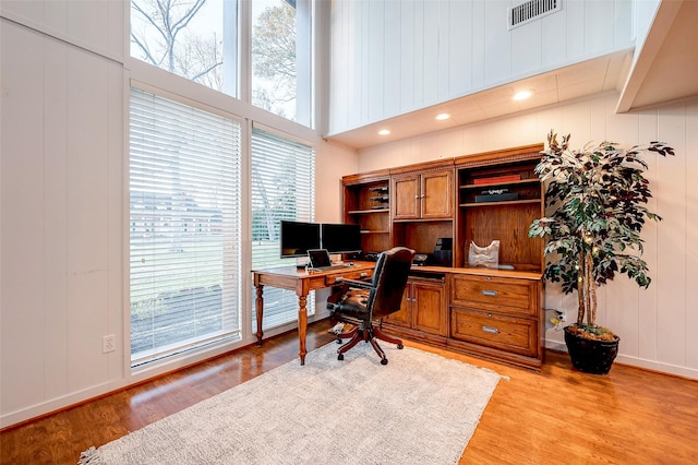 home office featuring built in desk, visible vents, light wood-style floors, a decorative wall, and a high ceiling