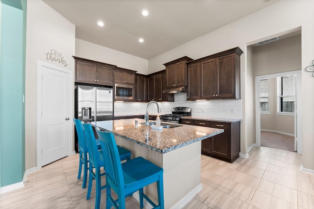 kitchen with dark brown cabinetry, an island with sink, appliances with stainless steel finishes, light stone counters, and a sink