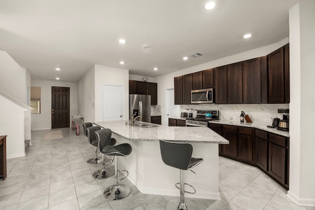 kitchen with a kitchen bar, stainless steel appliances, an island with sink, light stone countertops, and decorative backsplash