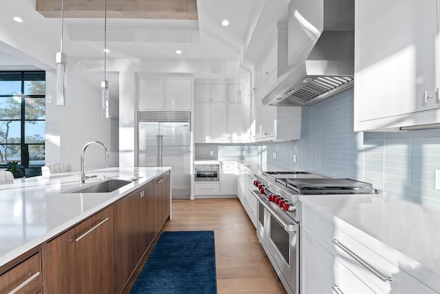 kitchen with sink, white cabinetry, wall chimney range hood, premium appliances, and hanging light fixtures