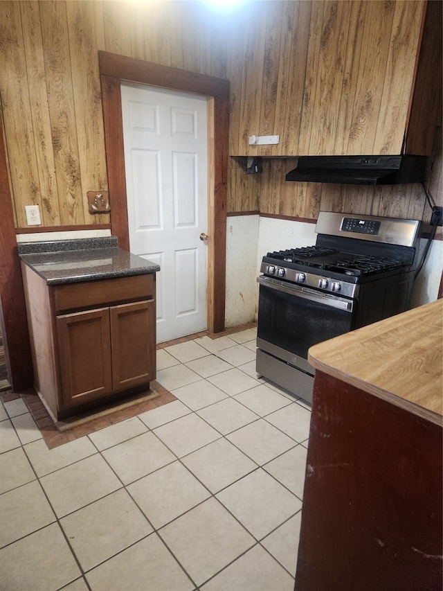 kitchen featuring stainless steel gas stove, light tile patterned floors, dark countertops, wood walls, and exhaust hood