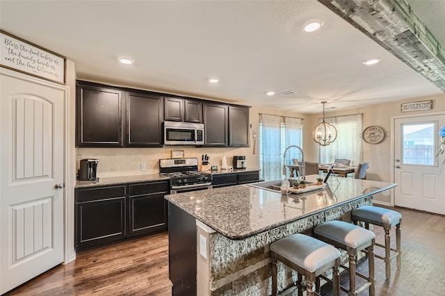 kitchen with stainless steel appliances, an island with sink, decorative light fixtures, and light stone countertops