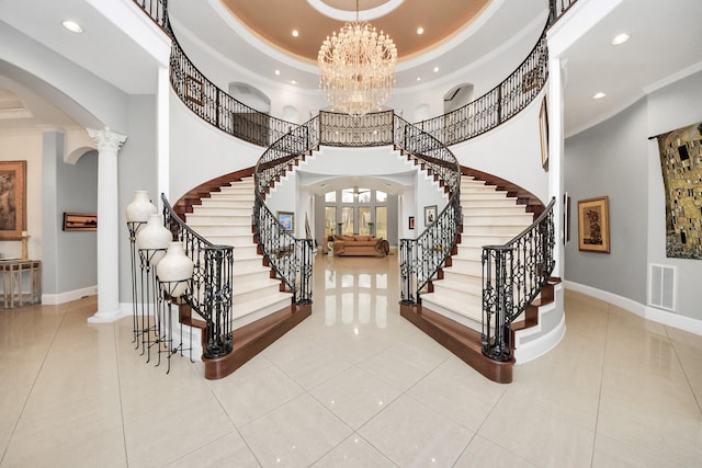 tiled foyer entrance featuring decorative columns, a towering ceiling, ornamental molding, a raised ceiling, and a chandelier