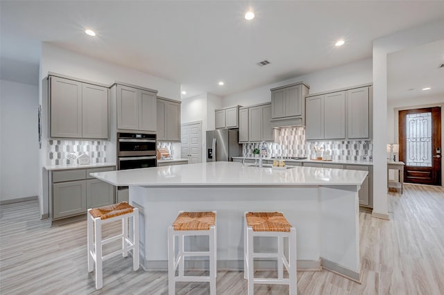 kitchen featuring appliances with stainless steel finishes, a breakfast bar, a kitchen island with sink, light countertops, and gray cabinetry