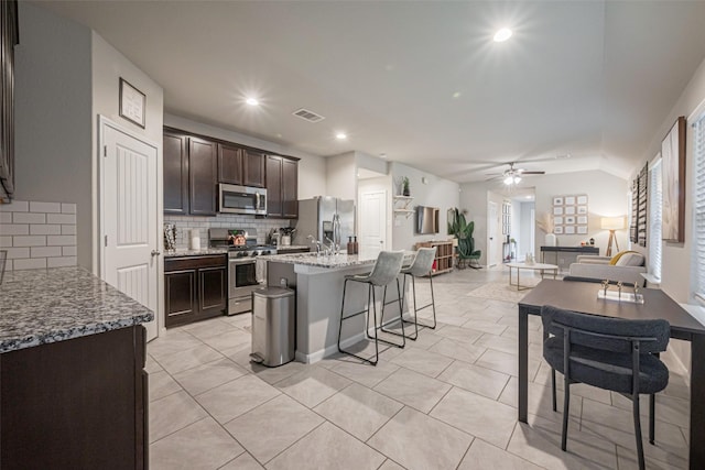 kitchen featuring a center island with sink, stainless steel appliances, ceiling fan, light stone counters, and a breakfast bar area