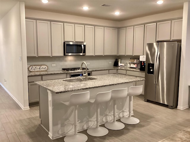 kitchen with an island with sink, light hardwood / wood-style flooring, light stone countertops, sink, and appliances with stainless steel finishes