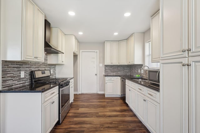 kitchen with tasteful backsplash, dark countertops, wall chimney exhaust hood, appliances with stainless steel finishes, and a sink