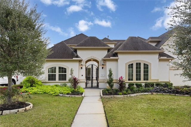 french country inspired facade with a shingled roof, a front yard, and stucco siding