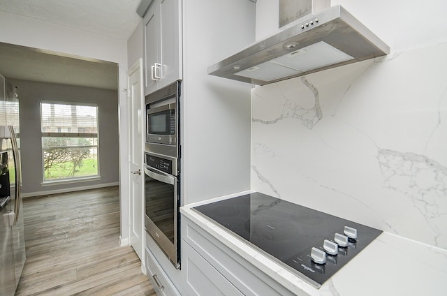 kitchen with light wood-style flooring, light stone counters, range hood, white cabinetry, and appliances with stainless steel finishes