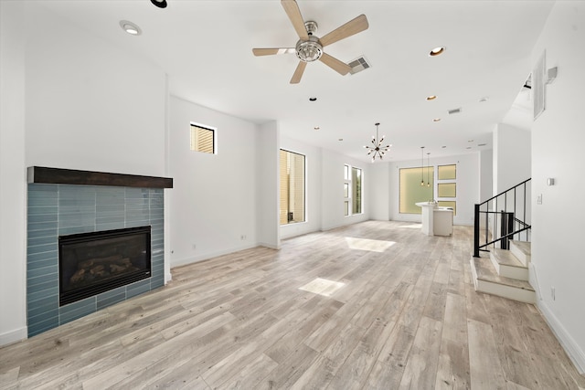 unfurnished living room featuring visible vents, a tiled fireplace, stairway, light wood-style floors, and ceiling fan with notable chandelier