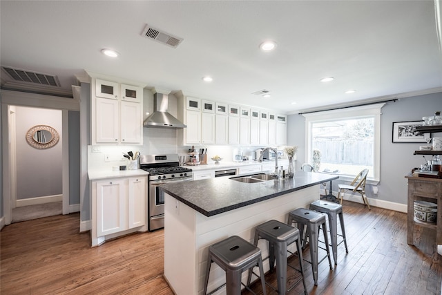 kitchen with wall chimney range hood, stainless steel range with gas cooktop, sink, white cabinetry, and a kitchen island with sink