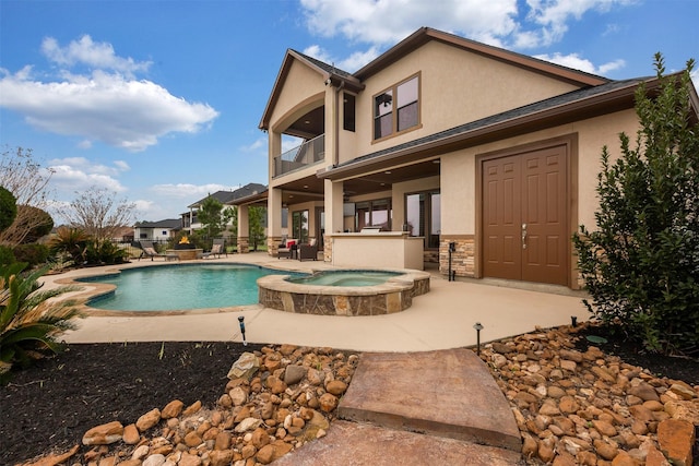 back of house featuring a patio, stone siding, a pool with connected hot tub, stucco siding, and a balcony