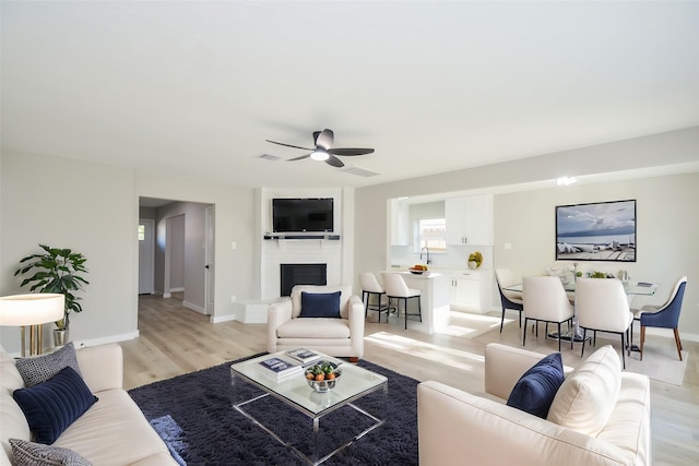 living area with baseboards, a ceiling fan, light wood finished floors, and a brick fireplace