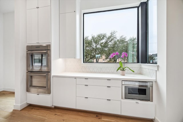 kitchen with light countertops, white cabinets, double oven, light wood-type flooring, and backsplash