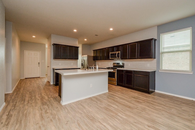 kitchen featuring a kitchen island with sink, light countertops, appliances with stainless steel finishes, light wood finished floors, and tasteful backsplash