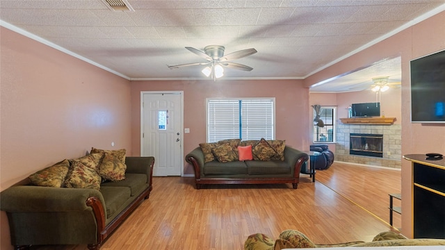 living area featuring crown molding, visible vents, light wood-style flooring, ceiling fan, and a stone fireplace