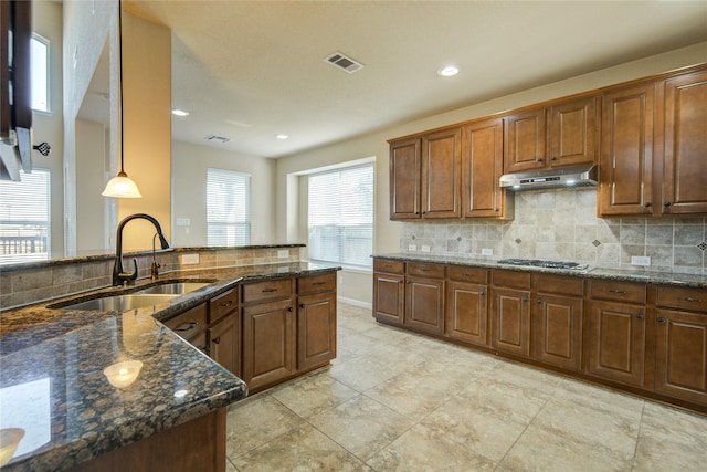 kitchen with under cabinet range hood, a sink, hanging light fixtures, decorative backsplash, and dark stone countertops