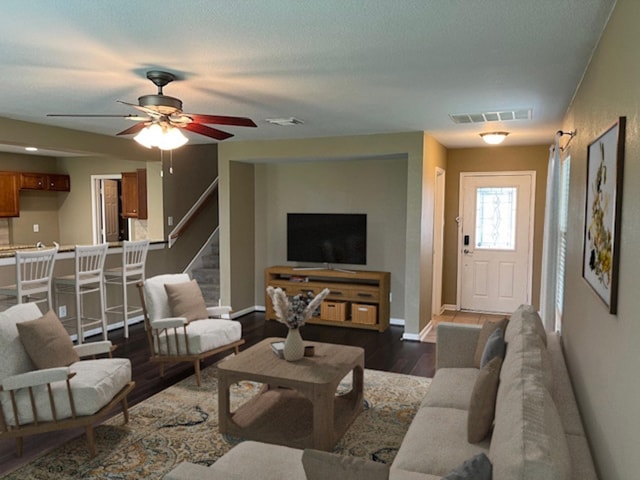 living room with dark wood-style flooring, a ceiling fan, visible vents, baseboards, and stairway