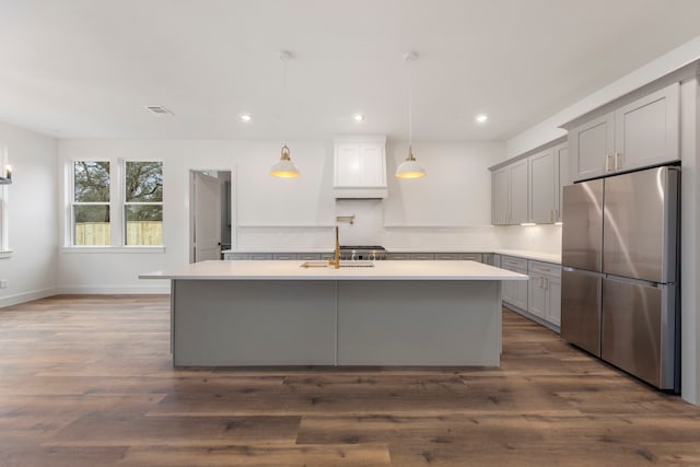 kitchen featuring freestanding refrigerator, light countertops, a center island with sink, and hanging light fixtures