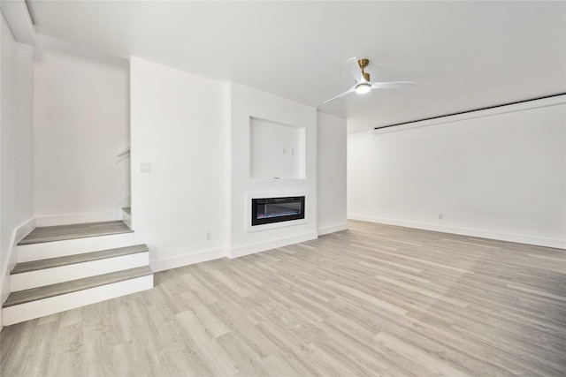 unfurnished living room with stairway, a ceiling fan, baseboards, light wood-style flooring, and a glass covered fireplace