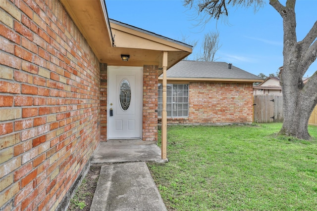 view of exterior entry with a shingled roof, fence, a lawn, and brick siding